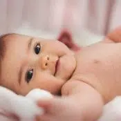 Un petit bébé qui sourit en regardant l'objectif. Il est couché sans vêtement sur une couverture blanche.
