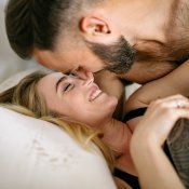 Un homme (brun avec barbe) et une femme (blonde aux cheveux longs) sont allongés dans un lit. L'homme est positionné au dessus de la femme. Ils se sourient et sont prêts à s'embrasser.