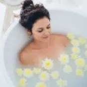 Une femme brune prend un bain contre l'eczéma. L'eau est trouble et à la surface flotte des fleurs jaunes.