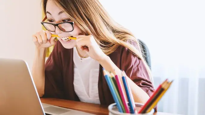 Stress au travail : une femme assise à son bureau devant son ordinateur avec un stylo dans la bouche, stressée et crispée. Rideau blanc en fond et pot de crayons/stylos en premier plan.